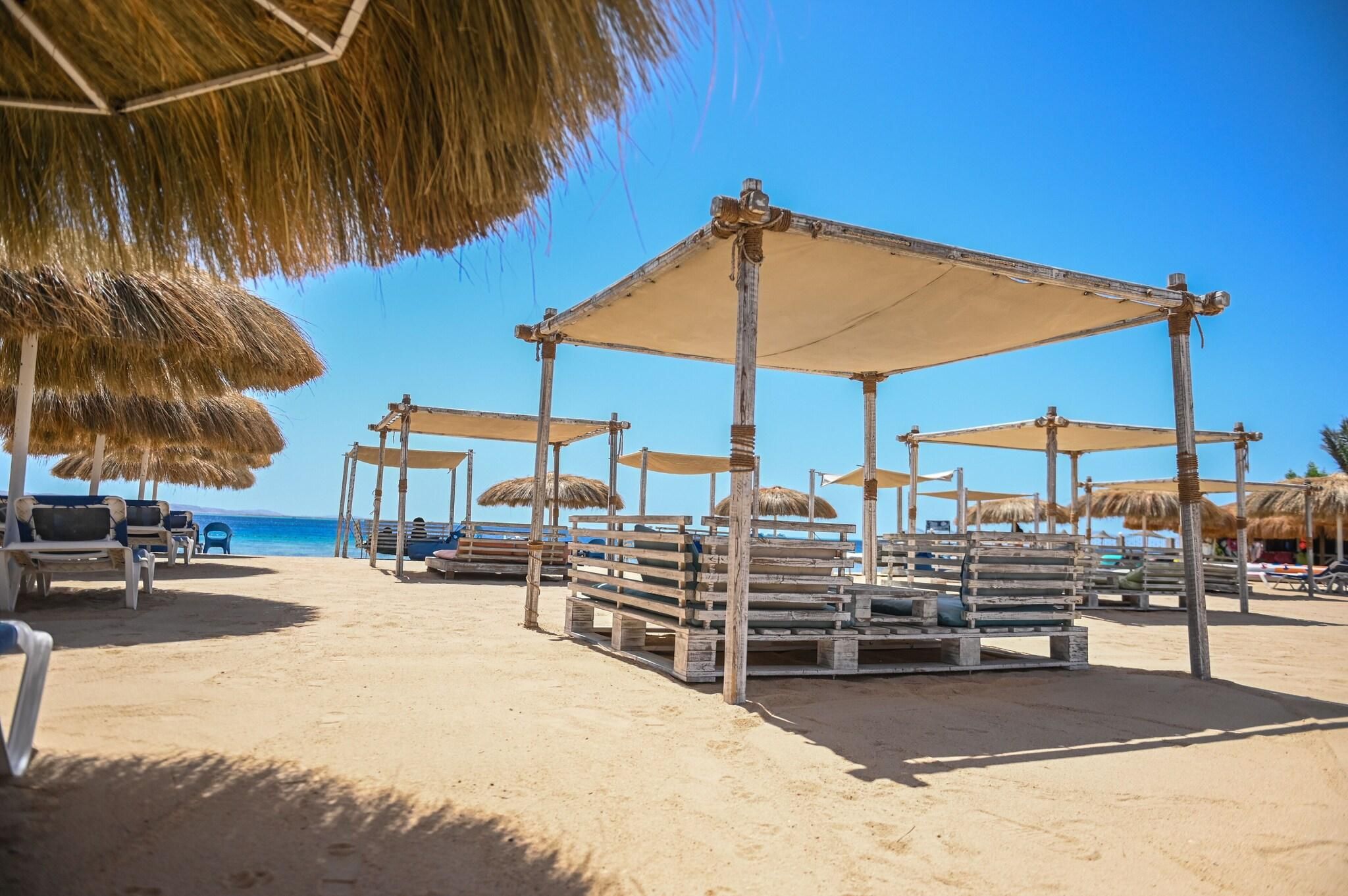 Ein Strand mit Holzbauten und Reetsonnenstühlen, die Schatten spenden.