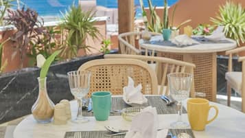 Un área de comedor al aire libre con vistas al mar, con mobiliario de mimbre, una mesa redonda y diversos utensilios para comer.