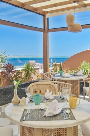 Área de refeições ao ar livre com vista para o mar, com móveis de vime, uma mesa redonda e diversos utensílios de jantar.