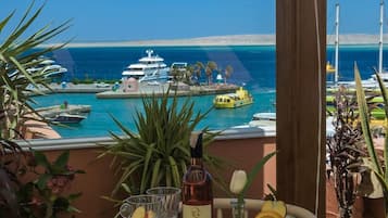 Una mesa servida con platos de comida, una botella de vino y una copa de bebida, con vistas a un puerto deportivo con barcos y un cielo azul despejado.