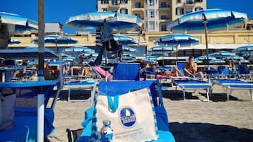 Una spiaggia con diversi lettini e ombrelloni, un edificio sullo sfondo e una borsa con un logo sulla sabbia.