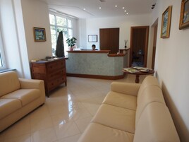 Una hall d'albergo con reception, un divano, un mobiletto in legno e una finestra con vista.