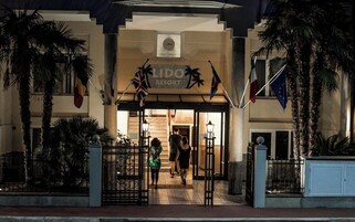 L'ingresso del Lido Resort, con un cartello sopra le porte, palme e bandiere.