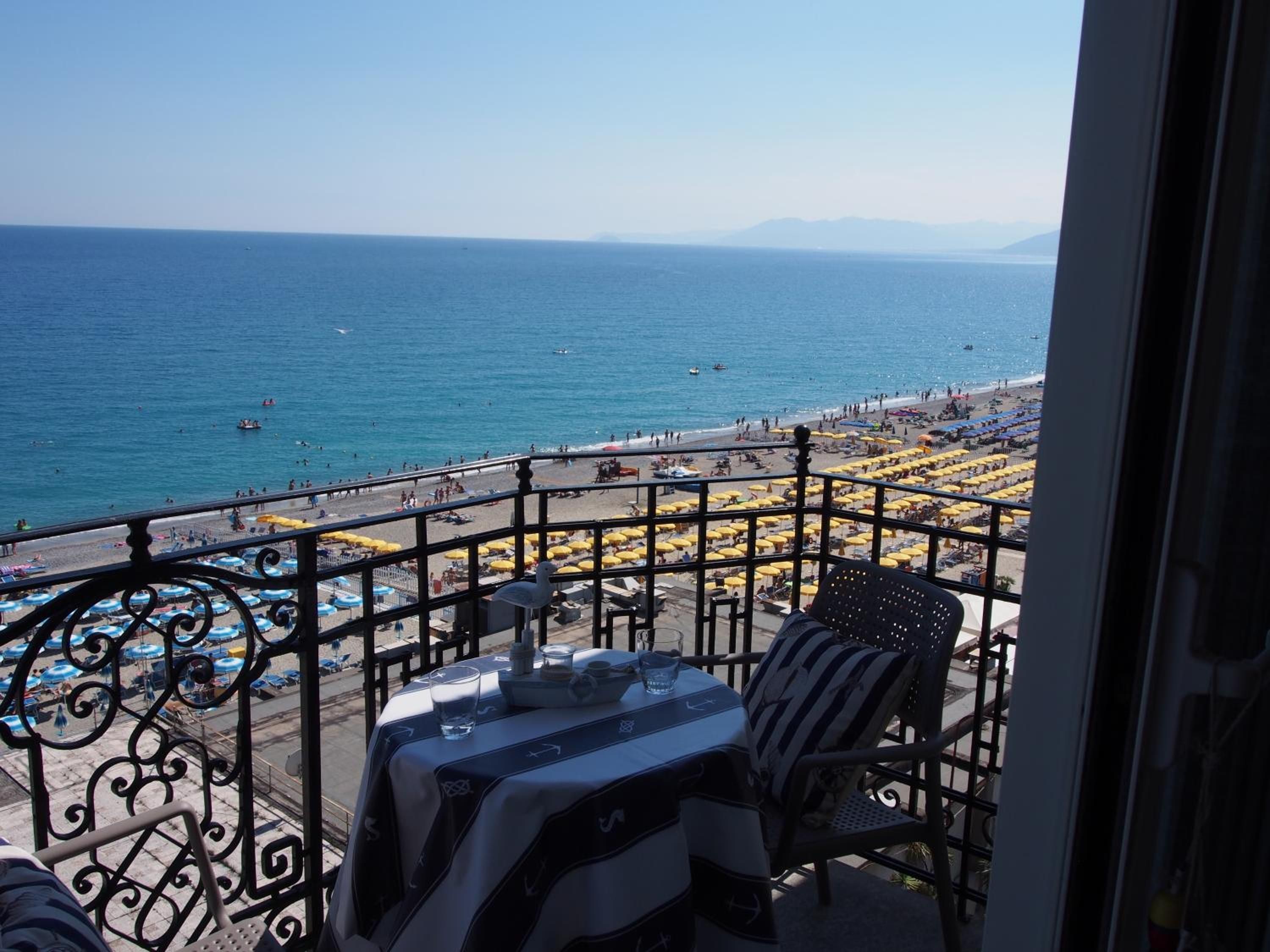 Un balcone con tavolo e sedie che si affaccia su una spiaggia con ombrelloni e persone.