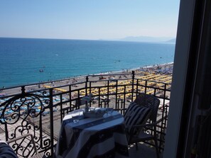A balcony with a table and chairs overlooking a beach with umbrellas and people. - Lido Resort (Finale Ligure)