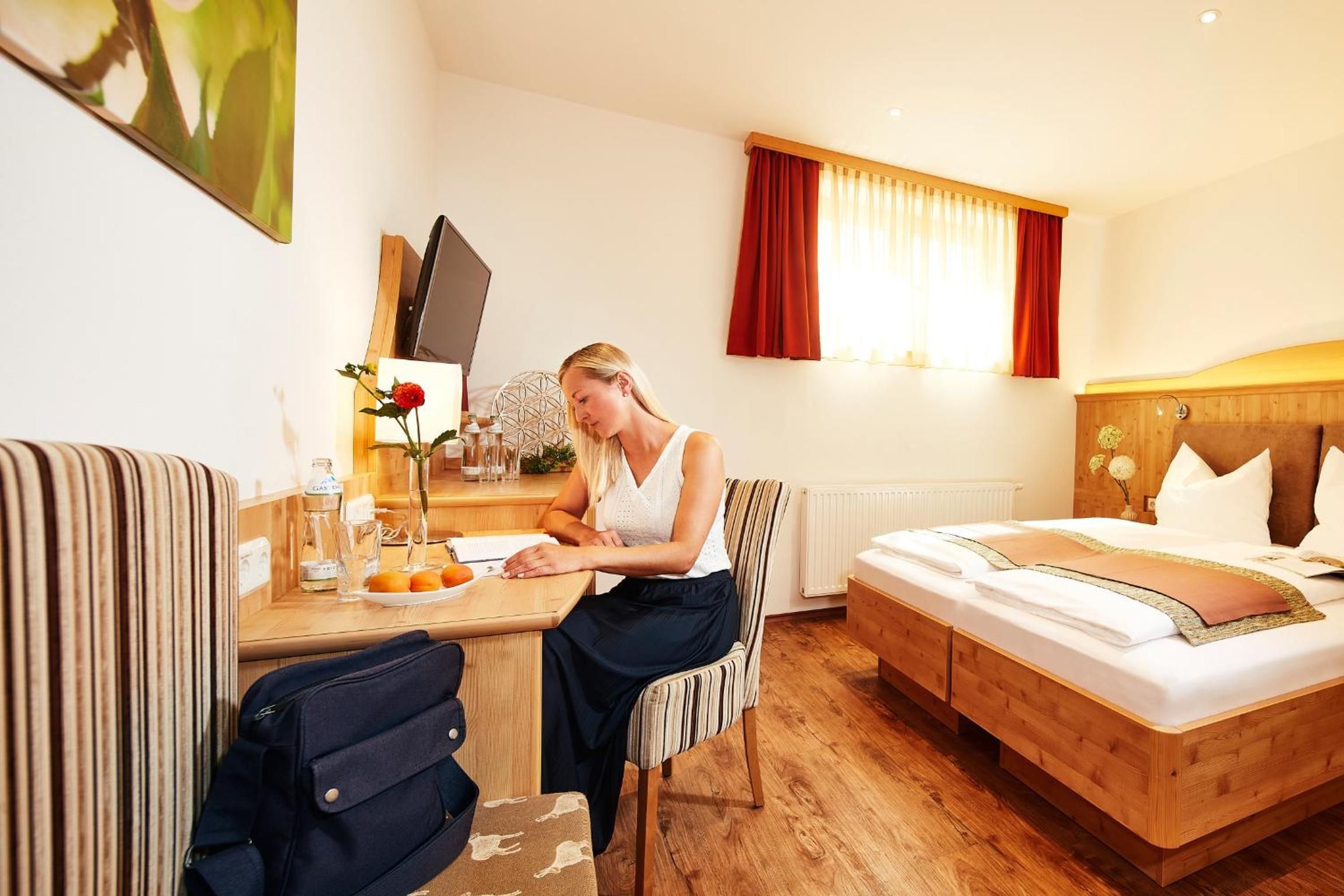 Ein Hotelzimmer mit einem Bett, einem Schreibtisch mit Stuhl, einem kleinen Tisch mit Vase und Obstteller sowie einem Fenster mit Vorhängen.