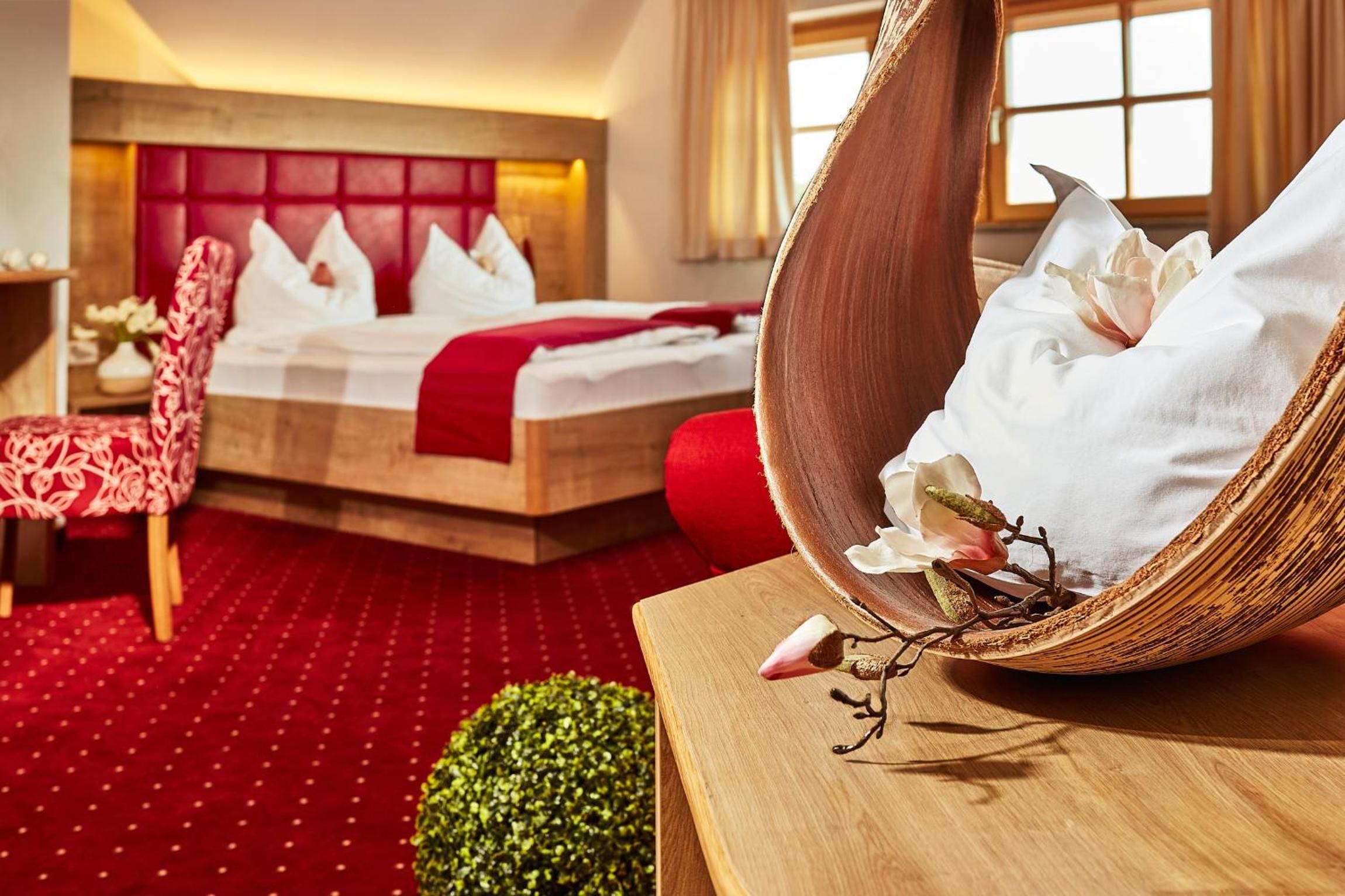 A hotel room with a red carpet, a bed with white linens, a red upholstered chair, and a wooden side table with a decorative item.