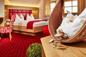 Habitación de hotel con alfombra roja, cama con ropa blanca, sillón tapizado en rojo y mesita de madera con un objeto decorativo.