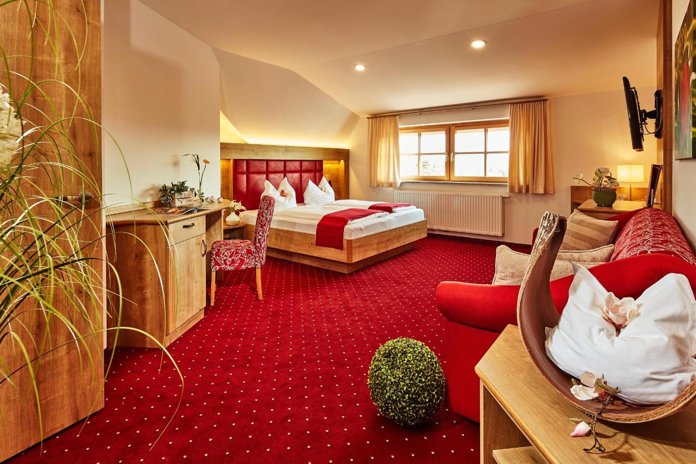 Ein Hotelzimmer mit rotem Teppich, einem Bett mit weißer Bettwäsche, einem Holztisch, einem roten Sofa und einer kleinen Zimmerpflanze.