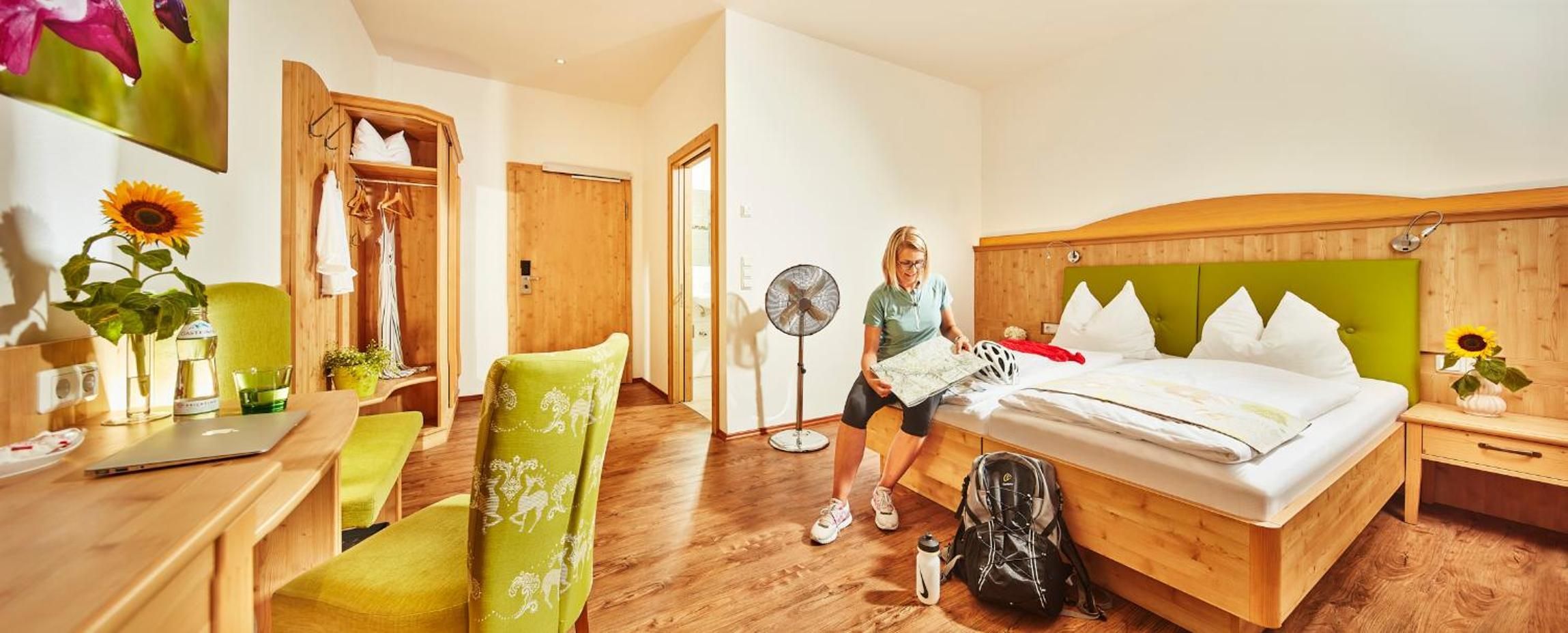A hotel room with a bed, a desk, a chair, a fan, and a suitcase.