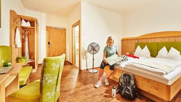 A hotel room with a bed, a desk, a chair, a fan, and a suitcase.