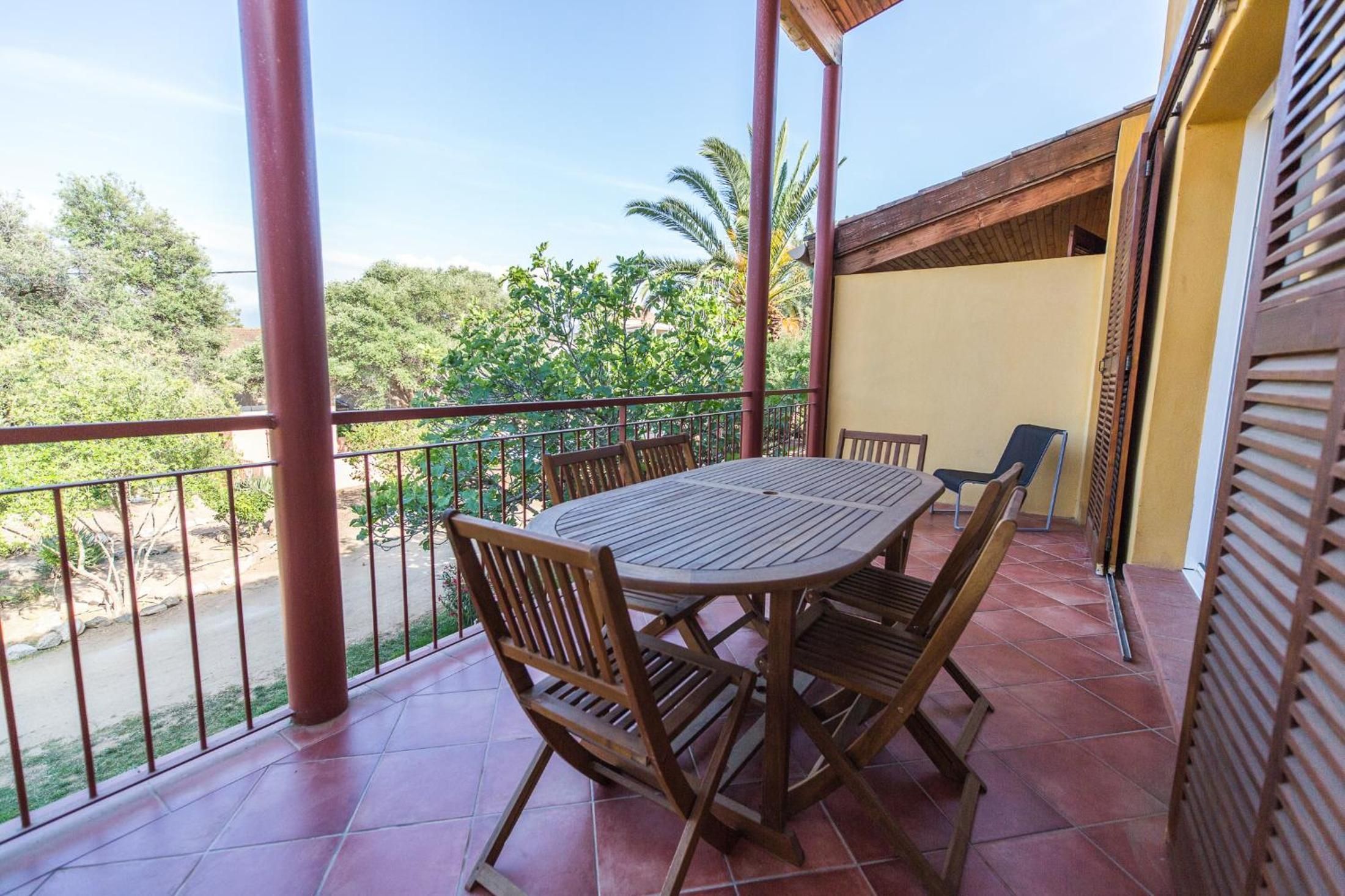 A covered outdoor patio with a round table and chairs, a railing, and a view of greenery and trees.
