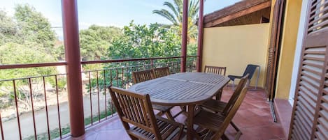 A covered outdoor patio with a round table and chairs, a railing, and a view of greenery and trees.