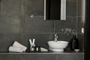 A modern bathroom with a white sink, dark tiles, a towel, toiletries, and a vase with flowers. - Aria Acropolis Suites (Athens)