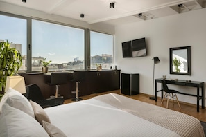 A modern hotel room with a large bed, a desk, a chair, a TV, and a view of the city. - Aria Acropolis Suites (Athens)