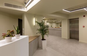 A modern reception area with a reception desk, potted plants, a wall-mounted light, and an elevator.