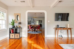 Ein modernes Wohnzimmer mit Holzboden, einem Essbereich mit rundem Tisch und Stühlen, einem an der Wand montierten Fernseher und einem Bücherregal mit Dekoration.