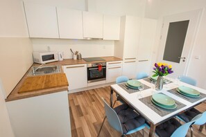 A modern kitchen with white cabinets, a microwave, a stove, and a sink. There is a dining area with a table set for two, chairs, and a vase with flowers. - Neklanova Apartments (Prague)