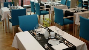 A restaurant with multiple tables set for dining, large windows offering a view of buildings, and modern pendant lighting. - Hotel Erwin Junker (Holice)