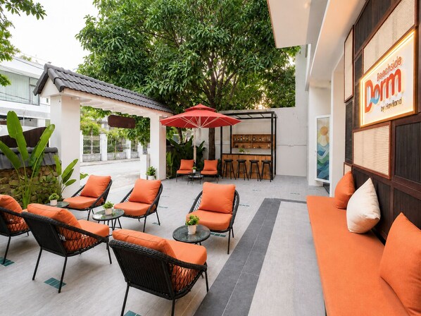 An outdoor seating area with wicker chairs and orange cushions, a red umbrella, and a bar area with stools. - Dorm Beachside Da Nang By Haviland (Da Nang)