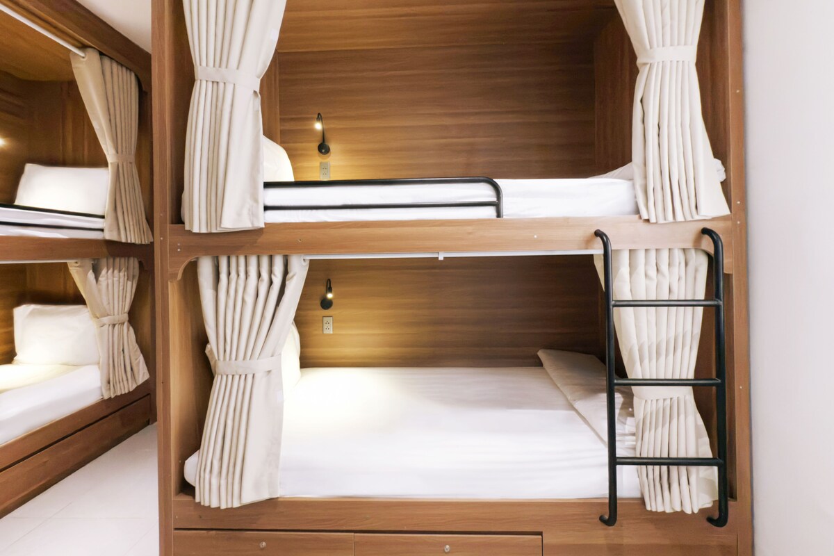 Une chambre avec un lit superposé, des rideaux, une table de chevet et une lampe fixée au mur.