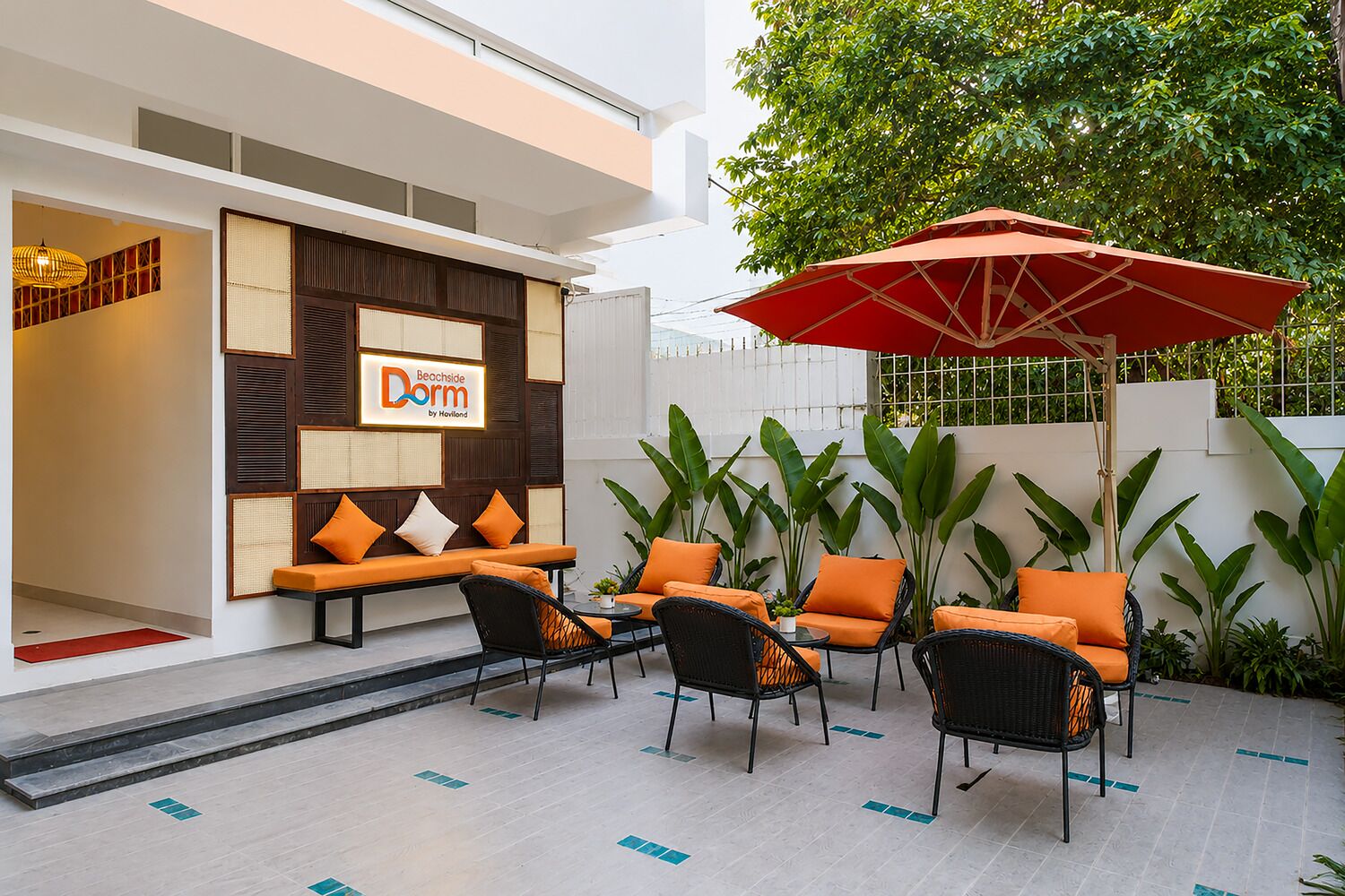 An outdoor seating area with wicker chairs and orange cushions, a red umbrella, and a sign for 'Beachside Dorm by Howell.'