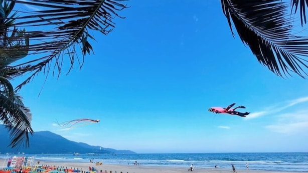 Una playa con sillones inflables de colores y una persona vestida de rosa volando una cometa.