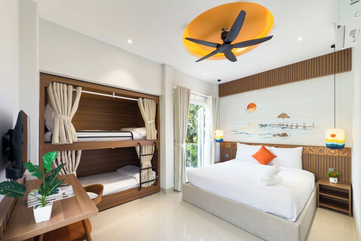 Une chambre d’hôtel avec un lit superposé, un bureau agrémenté d’une plante, un lit avec une literie blanche, un ventilateur de plafond et un tableau représentant une scène de plage.
