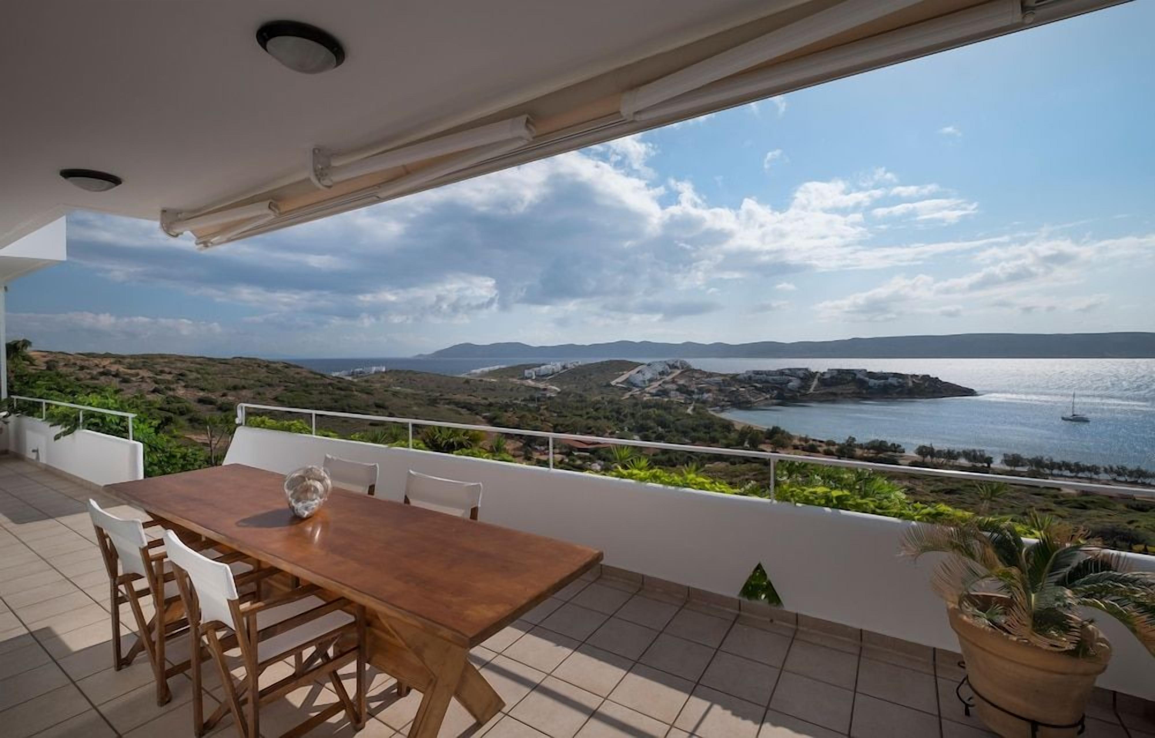 Una terraza con mesa y sillas de madera, con vistas a un paisaje costero con colinas y una masa de agua.