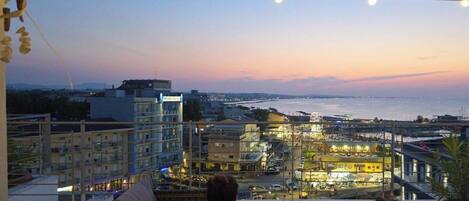 Uma varanda com vista para um horizonte urbano ao entardecer, incluindo edifícios e um corpo de água.