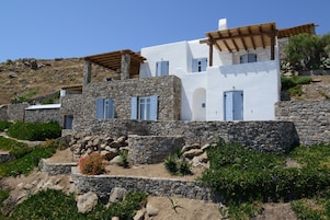 Un edificio blanco con muros de piedra y contraventanas azules, rodeado de follaje y rocas.