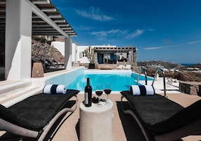 Un área junto a la piscina con sillones de descanso, una mesa con una botella y vasos, y vistas al mar y a las colinas.