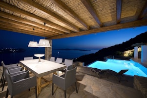 Un espacio de comedor al aire libre con mesa y sillas, piscina y vista al mar al anochecer.
