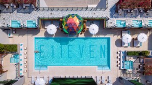 An aerial view of a swimming pool with a geometric pattern on the water's surface, surrounded by lounge chairs and umbrellas. - The E11EVEN Hotel & Residences (Miami)