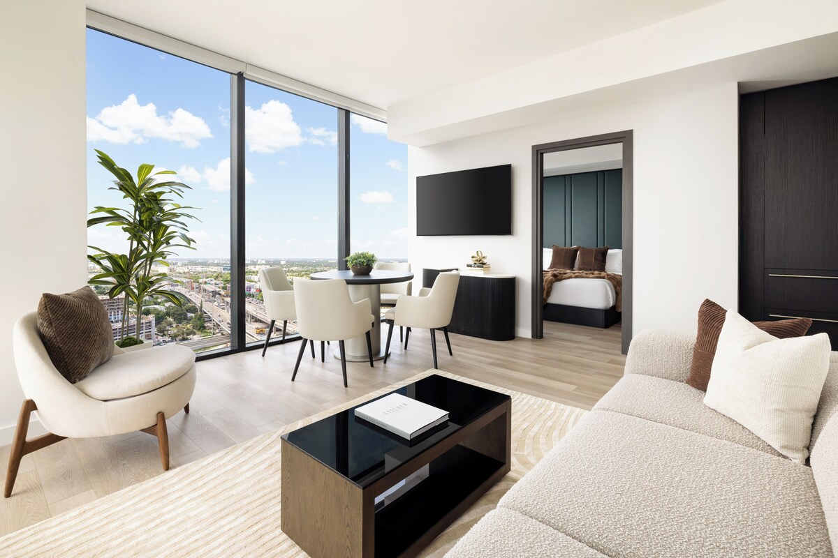 A modern living room with a large window, a flat-screen TV, a sofa, a coffee table, and a dining area with chairs.