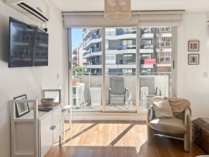 Un apartamento moderno con un amplio ventanal corredera que da a un balcón, un televisor de pantalla plana montado en la pared y una disposición de mobiliario minimalista.