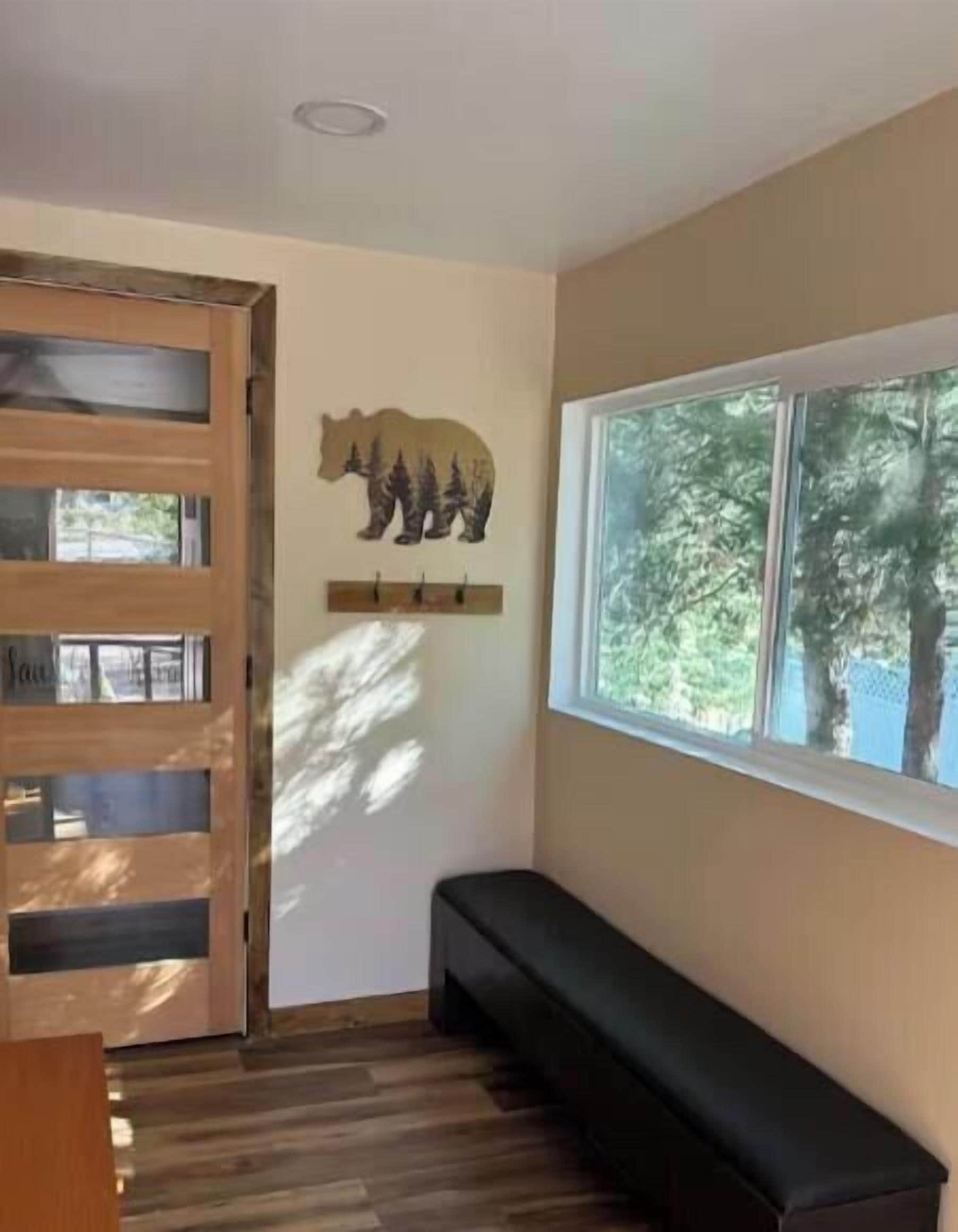 Una habitación con un banco de madera, un adorno de oso en la pared, una ventana con vistas a árboles y una puerta con paneles de vidrio.