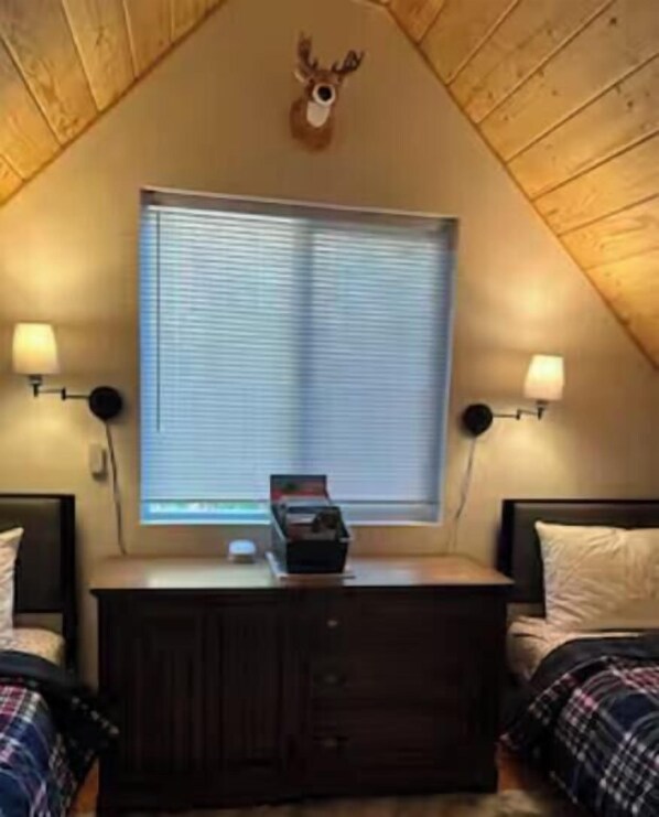 Una habitación con techo inclinado, una cama con cabecera, un aparador, un ventanal con persianas y dos lámparas fijadas a la pared.