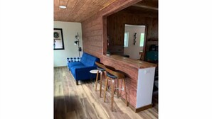 Una habitación con techo de madera, un sofá azul, una mesita, dos taburetes de bar y un electrodoméstico blanco.