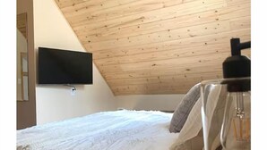 Un dormitorio con techo inclinado, una cama con ropa blanca, un televisor montado y una lámpara visible.