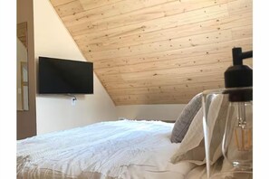 Ein Schlafzimmer mit schrägen Wänden, einem Bett mit weißer Bettwäsche, einem an der Wand befestigten Fernseher und einer sichtbaren Leuchte.