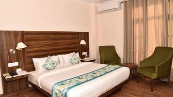 A hotel room with a large bed, two bedside tables, a green armchair, a small round table, and a window with curtains.