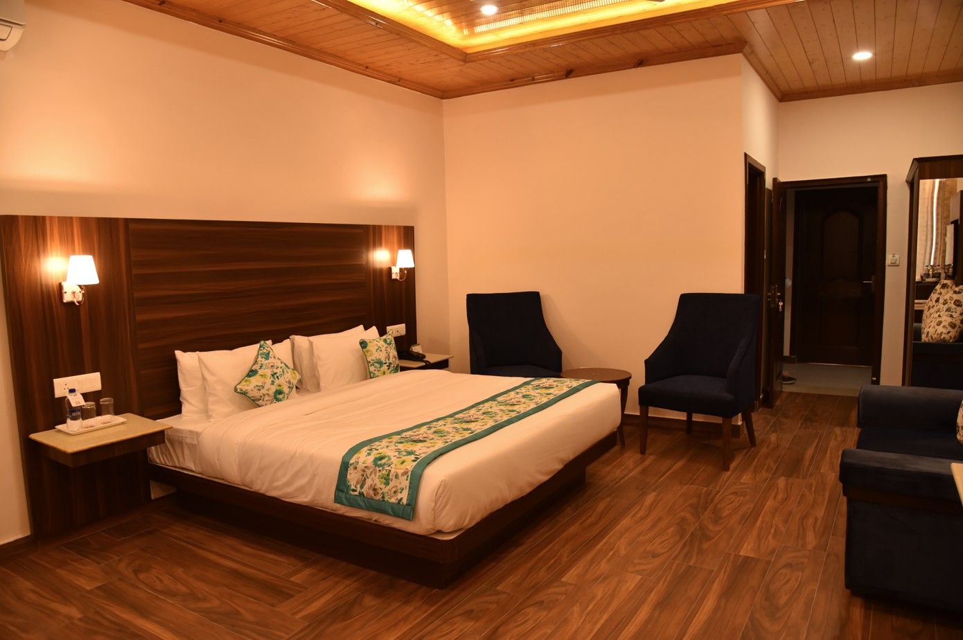 A hotel room with a wooden headboard, a bed with white linens and a green floral bedspread, two armchairs, a small table, and a nightstand with a lamp.