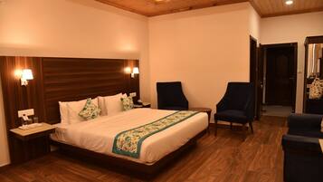 A hotel room with a wooden headboard, a bed with white linens and a green floral bedspread, two armchairs, a small table, and a nightstand with a lamp.
