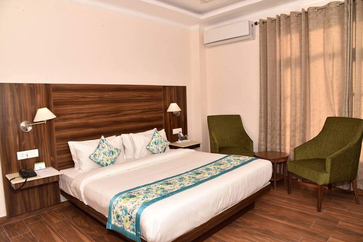 A hotel room with a large bed, two bedside tables, a green armchair, a small round table, and a window with curtains.
