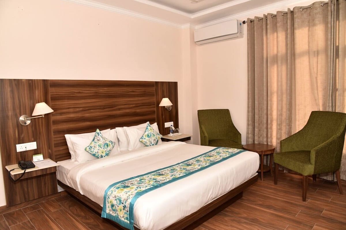 A hotel room with a large bed, two bedside tables, a green armchair, a small round table, and a window with curtains.