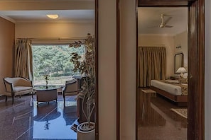 A hotel room with a bed, a desk, a chair, a potted plant, and a view of the outdoors. - Eagleton The Golf Resort (Ramanagara)