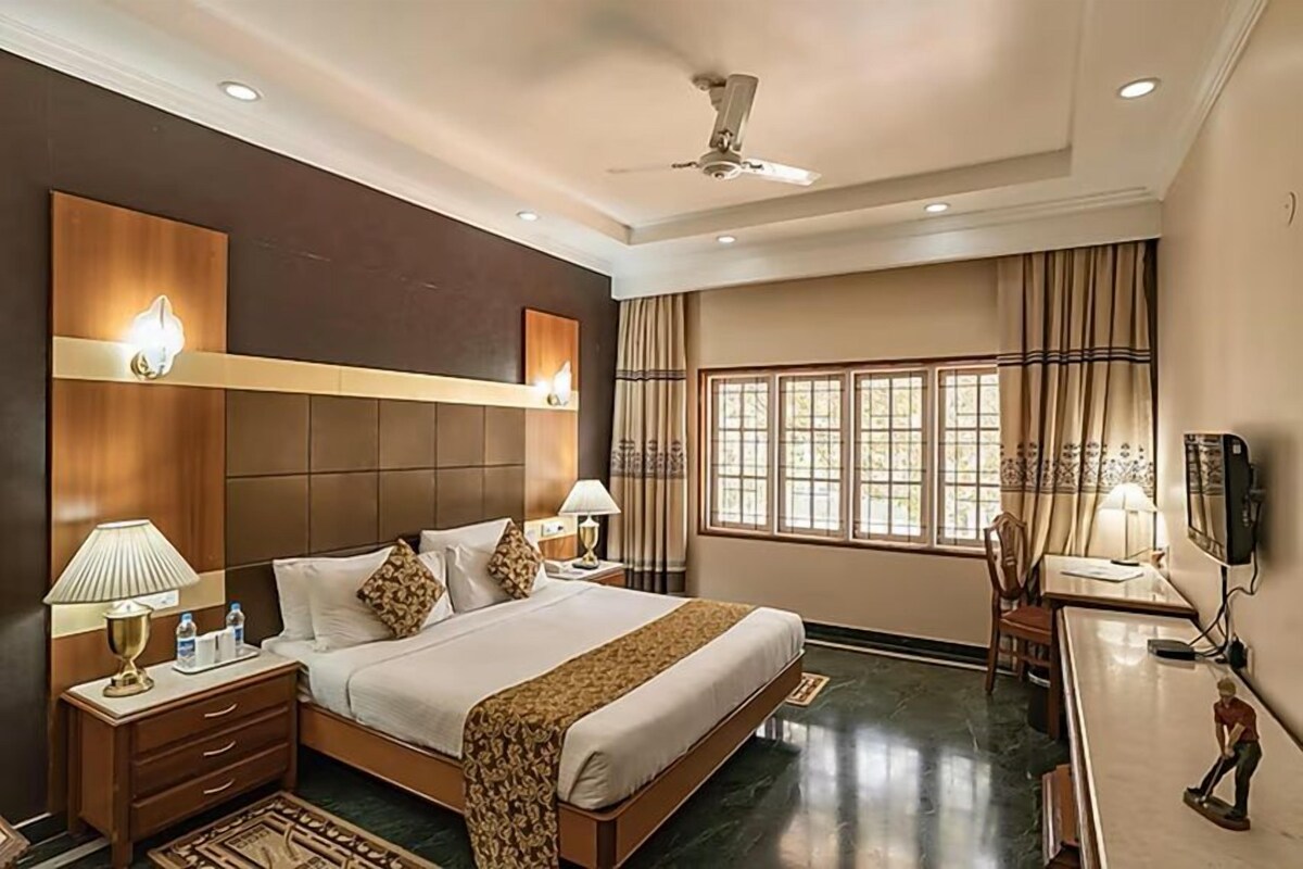 A hotel room with a large bed, bedside tables, a desk, a chair, a ceiling fan, and a window with curtains.