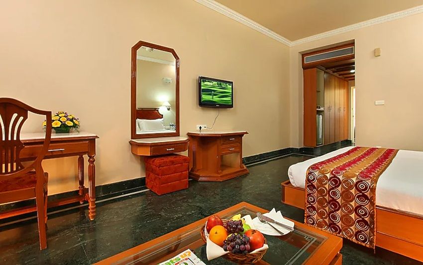 A hotel room with a bed, a desk with a chair, a vanity with a mirror, a television, and a fruit basket on a coffee table.
