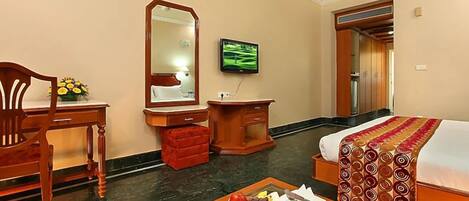 A hotel room with a bed, a desk with a chair, a vanity with a mirror, a television, and a fruit basket on a coffee table.